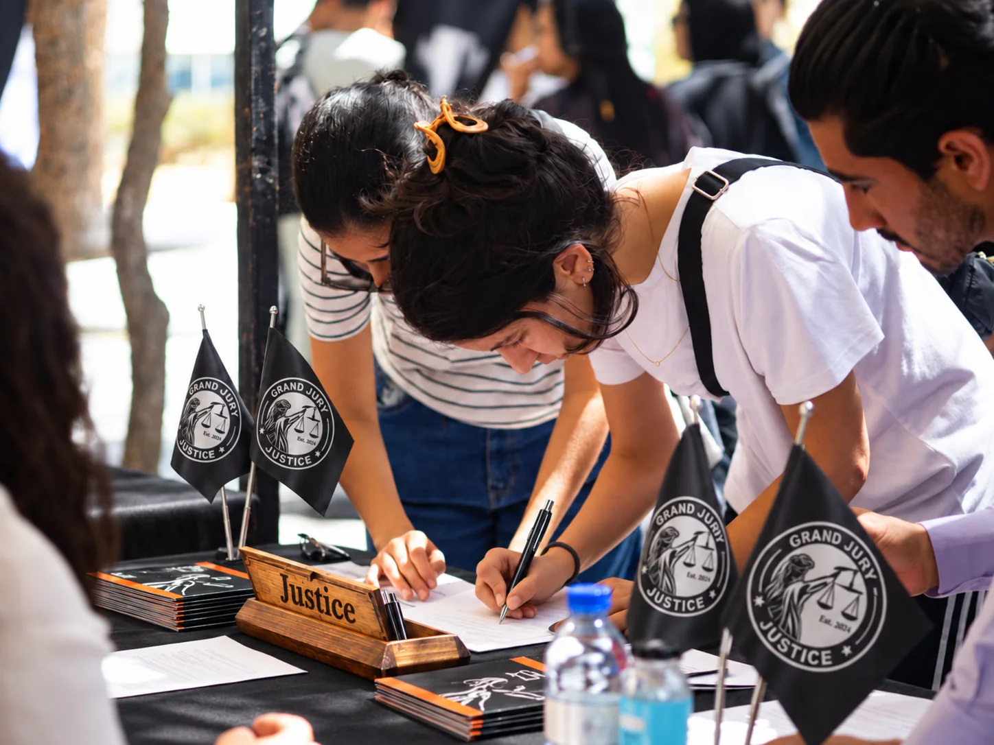 Illustrative scene of people reviewing and signing paperwork at a Grand Jury Justice-branded outdoor information table